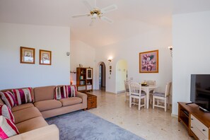 Un salon comprenant un canapé, une table basse, un meuble pour la télévision, une table à manger avec des chaises, une bibliothèque et un ventilateur de plafond.