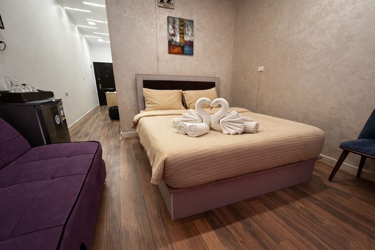 A modern hotel room with a large bed, a purple armchair, a desk with a computer, and a painting on the wall.