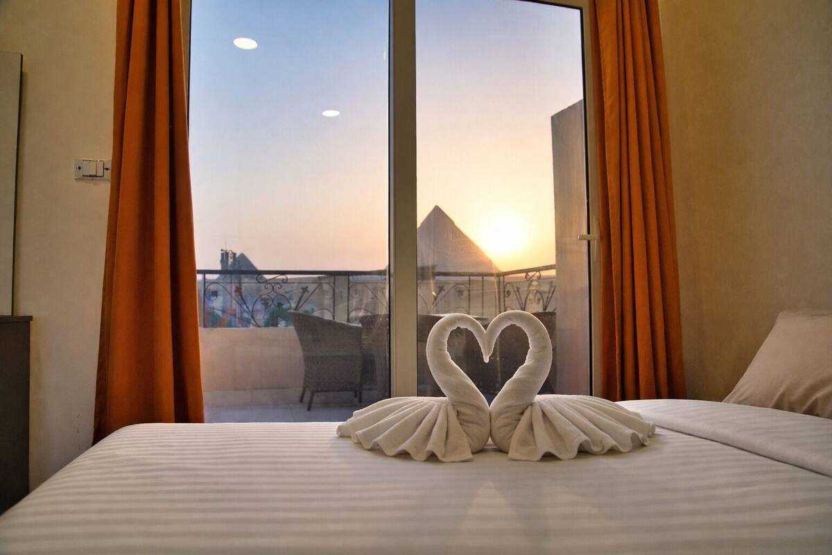 Une chambre d’hôtel avec un lit, une disposition de serviettes en forme de cœur et un balcon avec des rideaux orange.