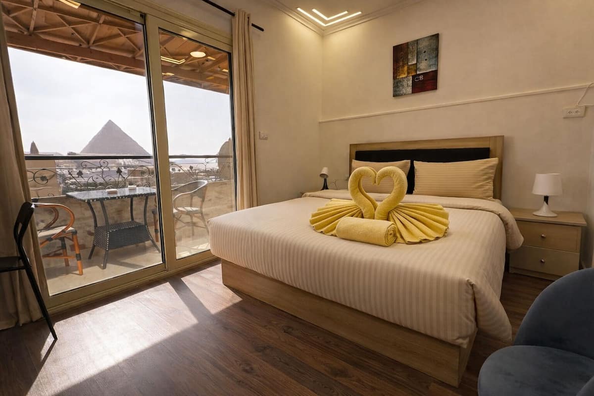 Une chambre d’hôtel avec un lit, une table de chevet, une chaise et une vue sur une pyramide, visible à travers une porte vitrée.