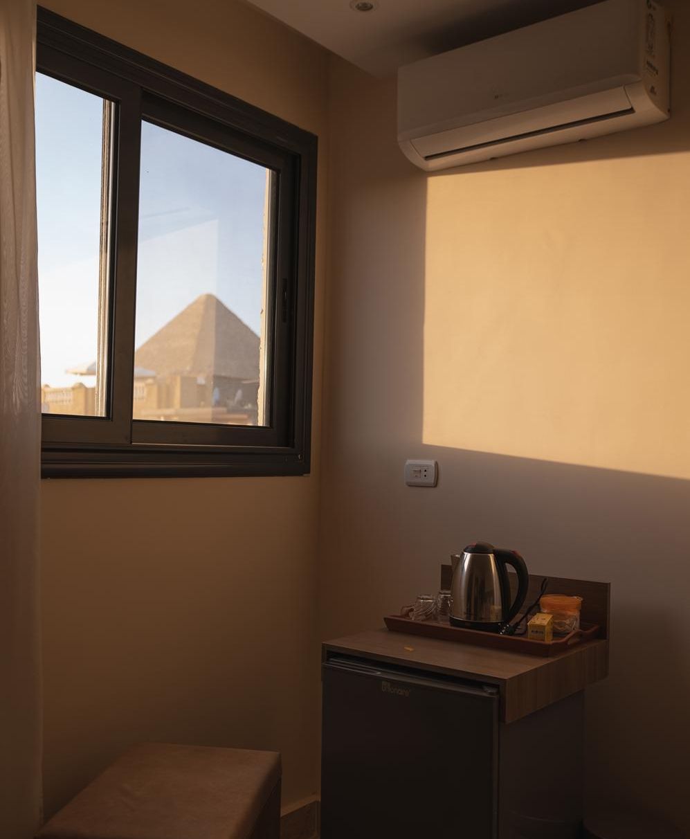 A compact kitchenette with a refrigerator, a kettle, and a coffee maker on a countertop. A window provides a view of a pyramid structure outside.