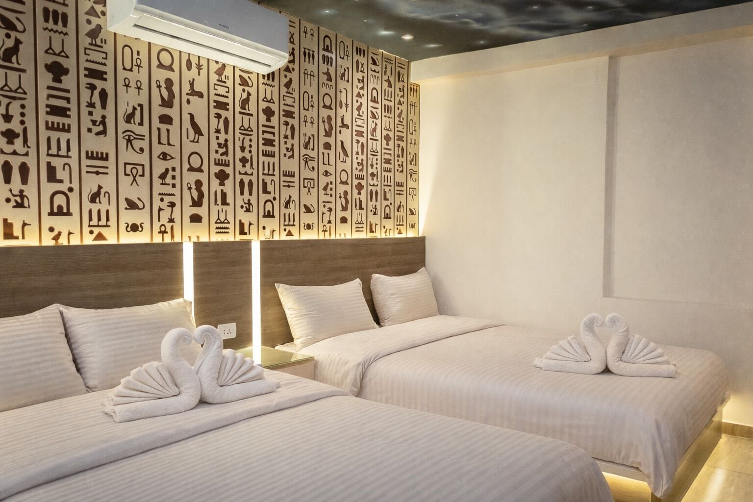 Habitación de hotel con dos camas, cada una adornada con una toalla con forma de cisne. Las paredes están decoradas con jeroglíficos.