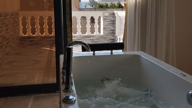 A modern bathroom with a bathtub, a shower, and a glass door leading to a balcony with a view of a pyramid and buildings. - Narmer Pyramids View (Giza)