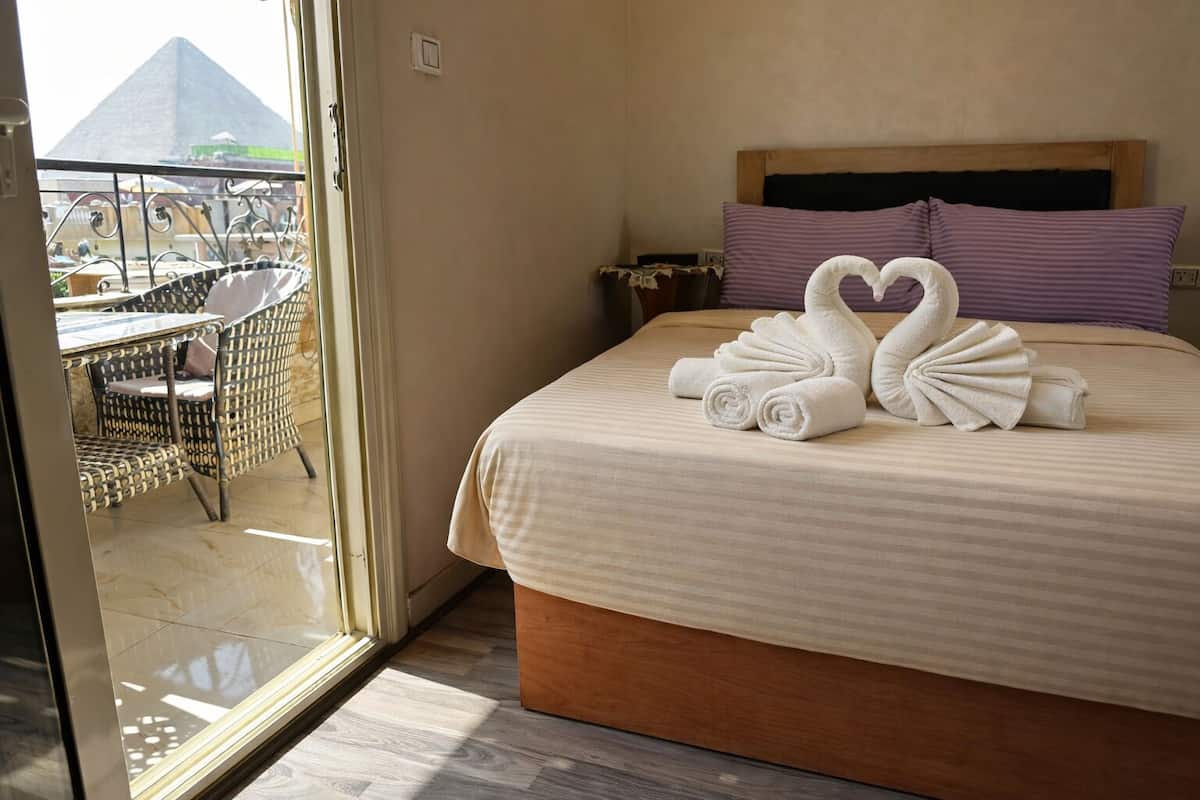 Une chambre d’hôtel avec un lit, deux serviettes en forme de cœur, une tête de lit en bois et une vue sur une pyramide, visible à travers une porte vitrée.