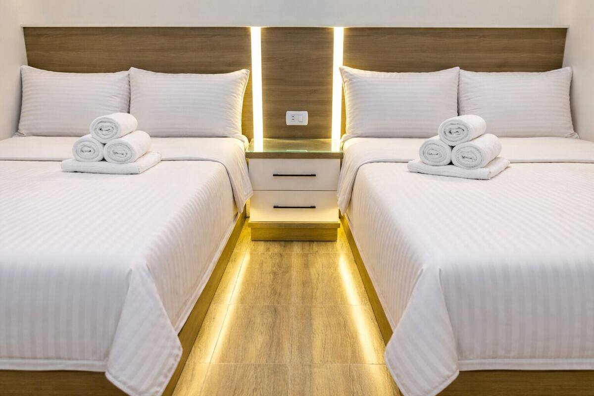 Une chambre d’hôtel avec deux lits, chacun équipé de draps blancs et de serviettes pliées. On y trouve une tête de lit en bois, une table de chevet avec une lampe et un petit meuble à tiroirs.