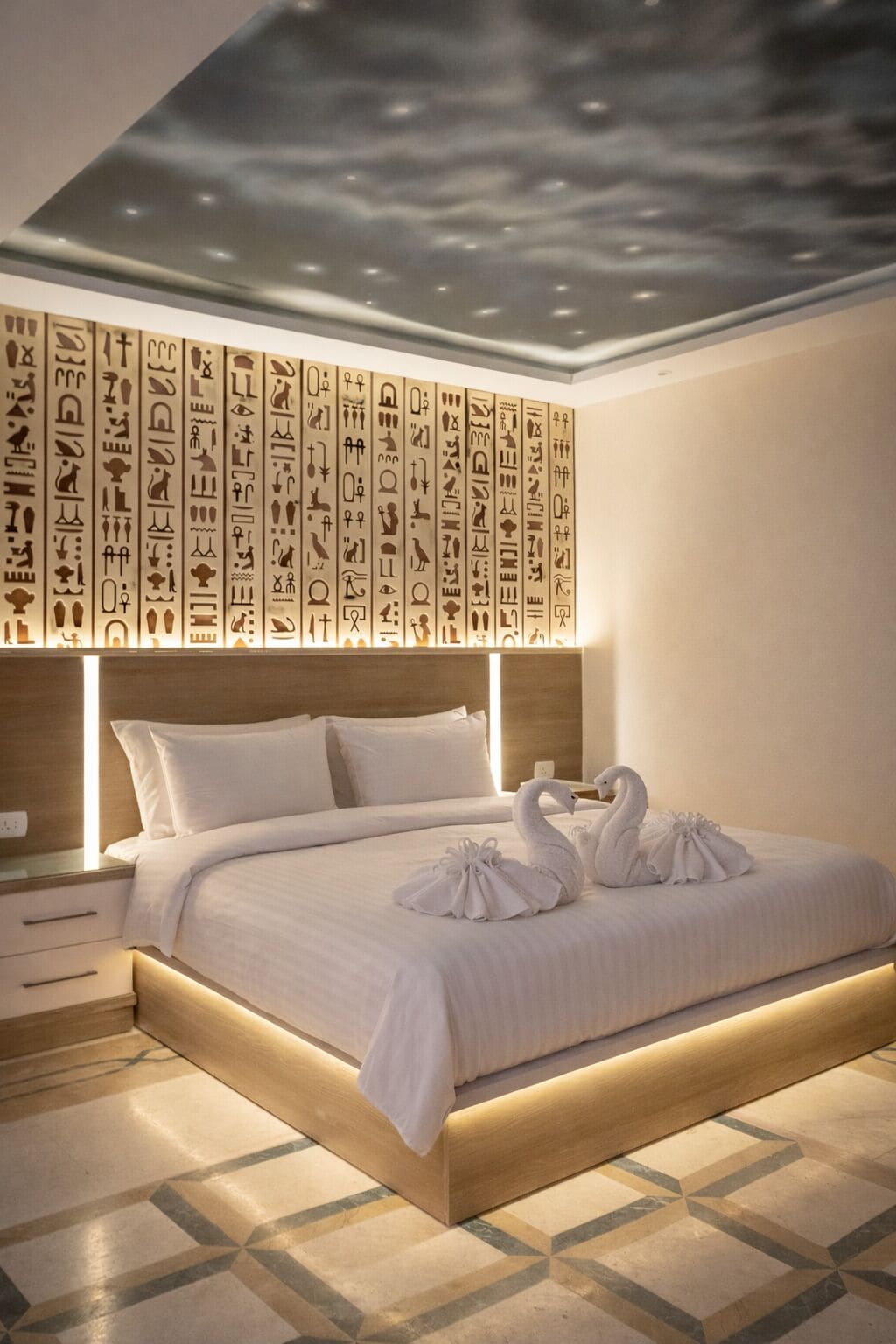 A modern hotel room with a large bed, a wooden headboard, and a ceiling with hieroglyphs.