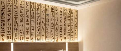 A modern hotel room with a large bed, a wooden headboard, and a ceiling with hieroglyphs.
