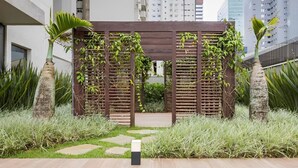 Un jardin extérieur moderne agrémenté d’une pergola en bois, de palmiers et d’un chemin menant à l’entrée d’un bâtiment.
