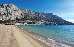 Ein Strand mit einem felsigen Berg im Hintergrund, klarem blauen Wasser und einer Reihe von Gebäuden entlang der Küste.