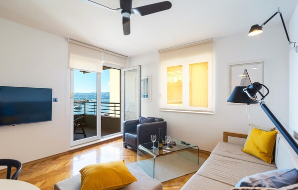 Ein modernes Wohnzimmer mit Flachbildfernseher, Deckenventilator, Glastisch und einem Sofa mit gelbem Kissen.