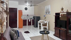 A living room with a sofa, a coffee table, a TV stand, a dining area with a table and chairs, a kitchen with cabinets and a refrigerator, and a ceiling fan.