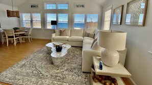 Ein geräumiges Wohnzimmer mit großem Fenster, Deckenventilator, Essbereich mit Tisch und Stühlen sowie eine gemütliche Sitzecke mit Sofa und Couchtisch.