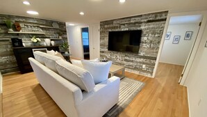 Ein modernes Wohnzimmer mit einem weißen Sofa, einem Flachbildfernseher an einer Wand mit Steinoptik und einer Kaffeemaschine auf einem Holzregal.