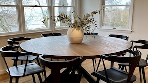 Ein runder Holztisch mit acht schwarzen Stühlen, eine weiße Vase mit Grünpflanzen und große Fenster mit Blick auf Bäume und Gebäude.