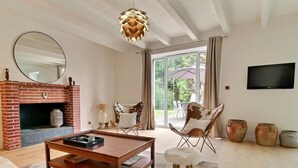 A living room with a brick fireplace, a round mirror, a wooden coffee table, a leather armchair, a sofa, and a television on the wall.
