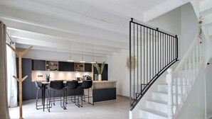 A modern kitchen with a central island, bar stools, pendant lighting, and a staircase with a black railing.