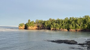 Ett kustlandskap med en stenig strand, stilla vatten och en skogbeklädd klippa.