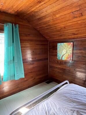 A room with wooden walls, a bed, a window with a painting, and a teal curtain.