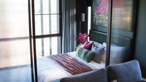 Un dormitorio con un ventanal grande, una cama con muchos almohadones, una mesita de noche con lámpara y una obra de arte colgada en la pared.