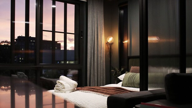 Una habitación de hotel moderna con un ventanal grande, una cama con toallas ordenadas, una lámpara de noche y vistas a la ciudad al atardecer.