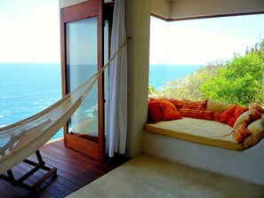 A hammock chair on a wooden deck overlooking the sea, a bed with orange and beige bedding, and a view of the ocean and greenery.
