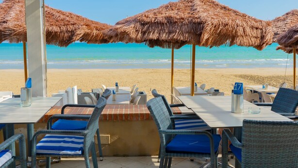 Ein Restaurant direkt am Strand, mit reetgedeckten Sonnenschirmen, Außenplätzen und Blick auf das Meer.