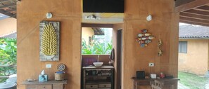 A room with a wooden table, a wall-mounted shelf with books, a small sink, and a window with a view of greenery. - Camburi Chalés (São Sebastião)
