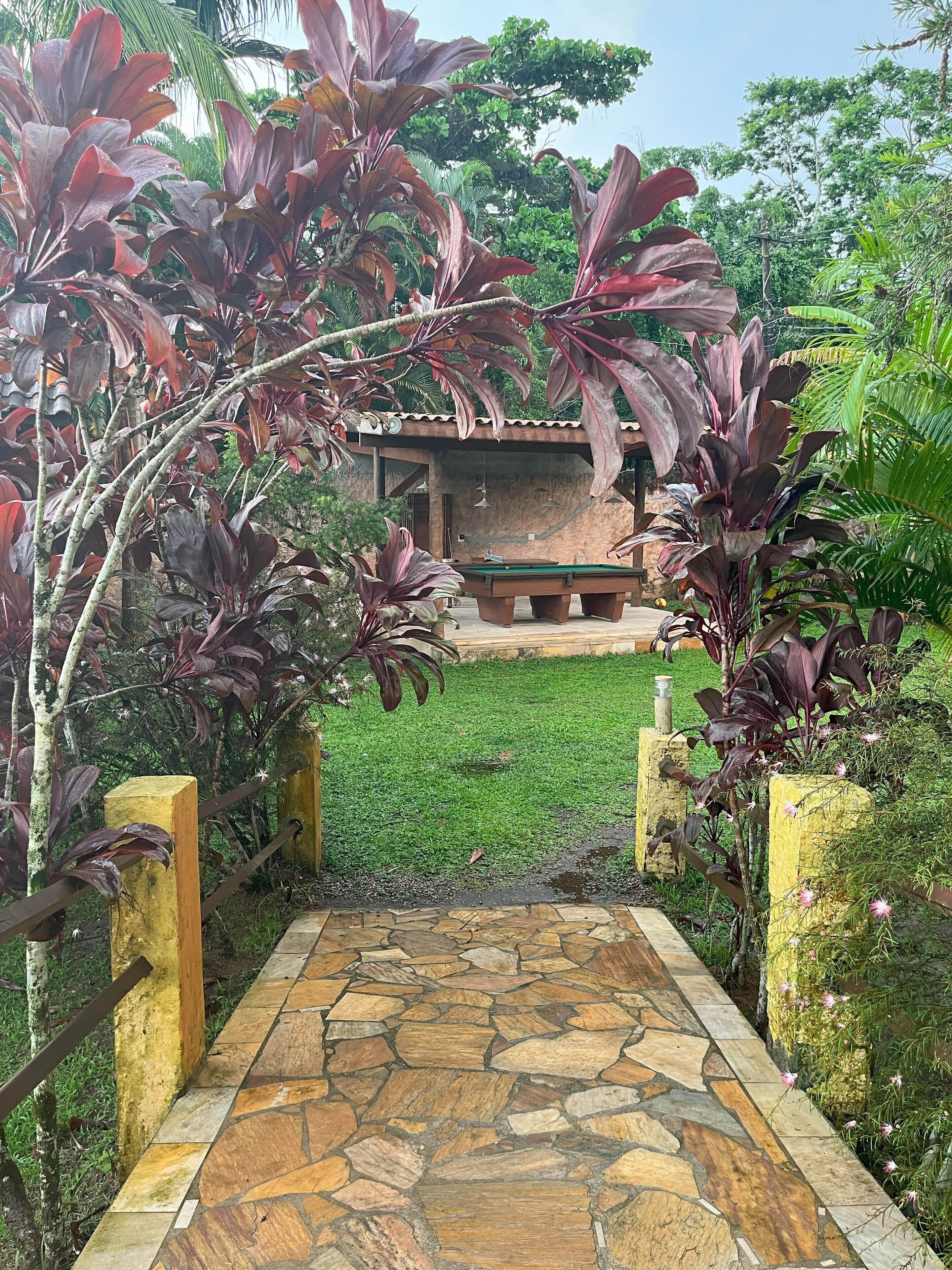 A stone pathway leading to a pool table in a garden with lush greenery and a small building in the background.