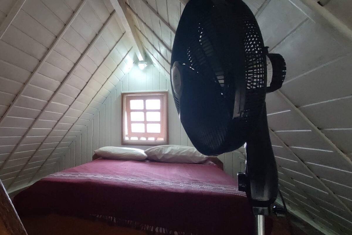 A small, dimly lit attic room with a sloped ceiling, a single bed with a red bedspread, a standing fan, and a window with a grid pattern.
