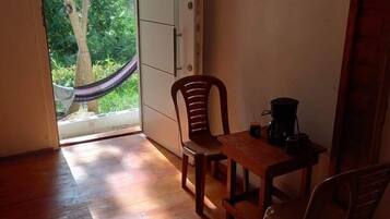 Una habitación pequeña con piso de madera, una mesa y sillas, una hamaca fuera de la puerta y vista a zonas verdes.