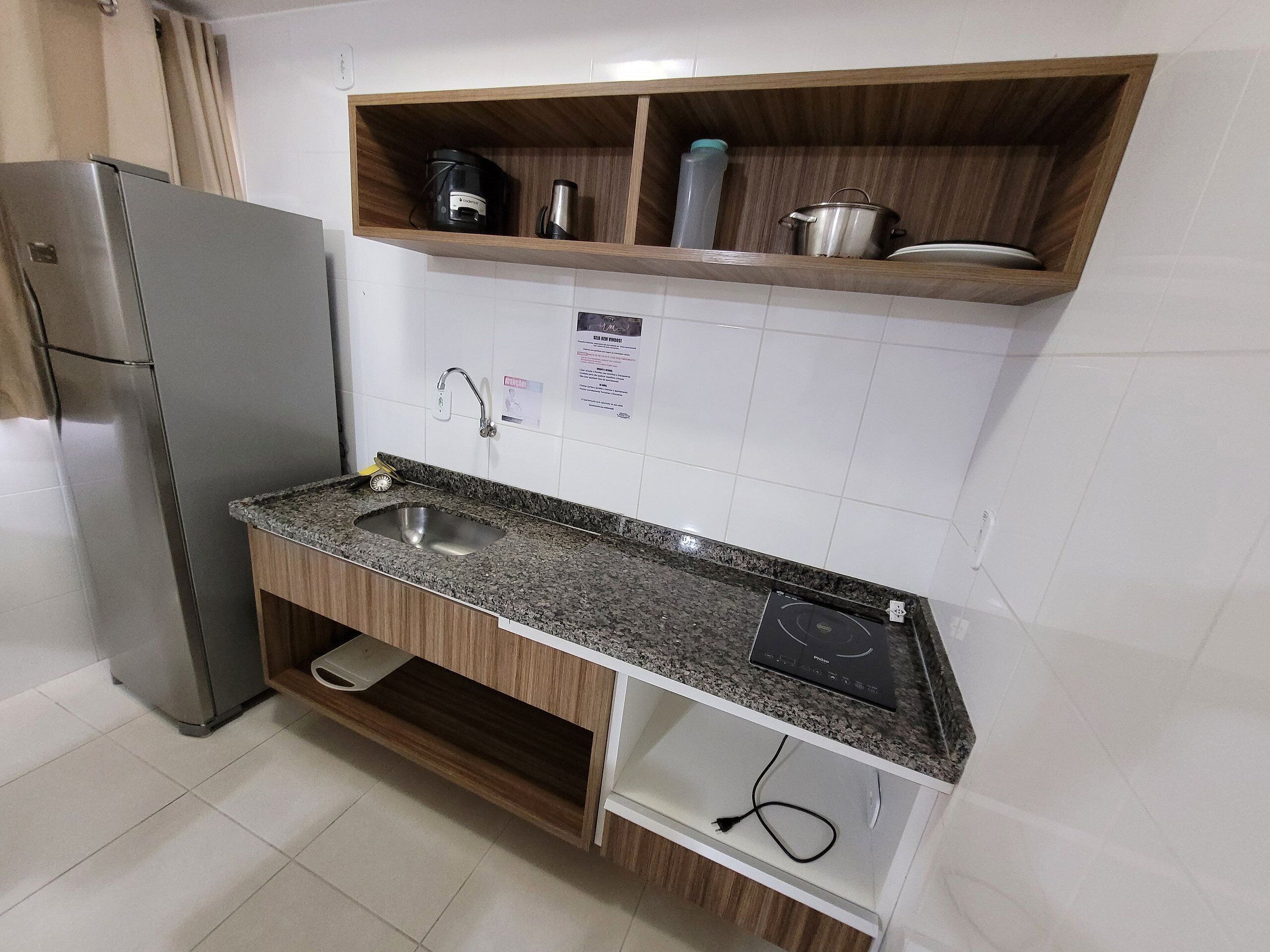 A kitchen with a stainless steel refrigerator, granite countertop, wooden cabinets, a built-in oven, and a shelf with kitchen appliances.