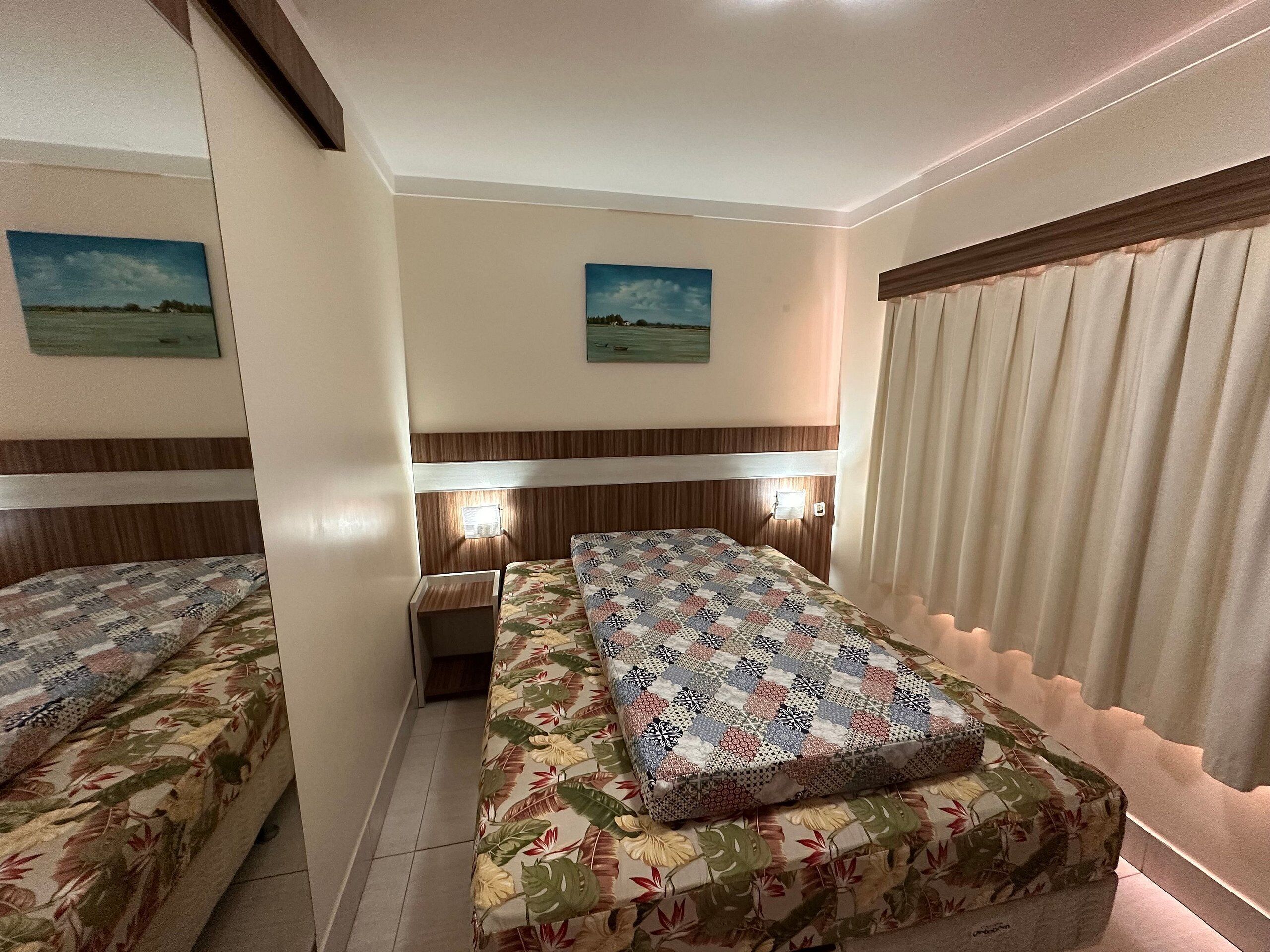 Hotellihuone, jossa on sänky, yöpöytä, seinään kiinnitetty lamppu, ikkuna verhoilla ja kaksi taulua seinällä.