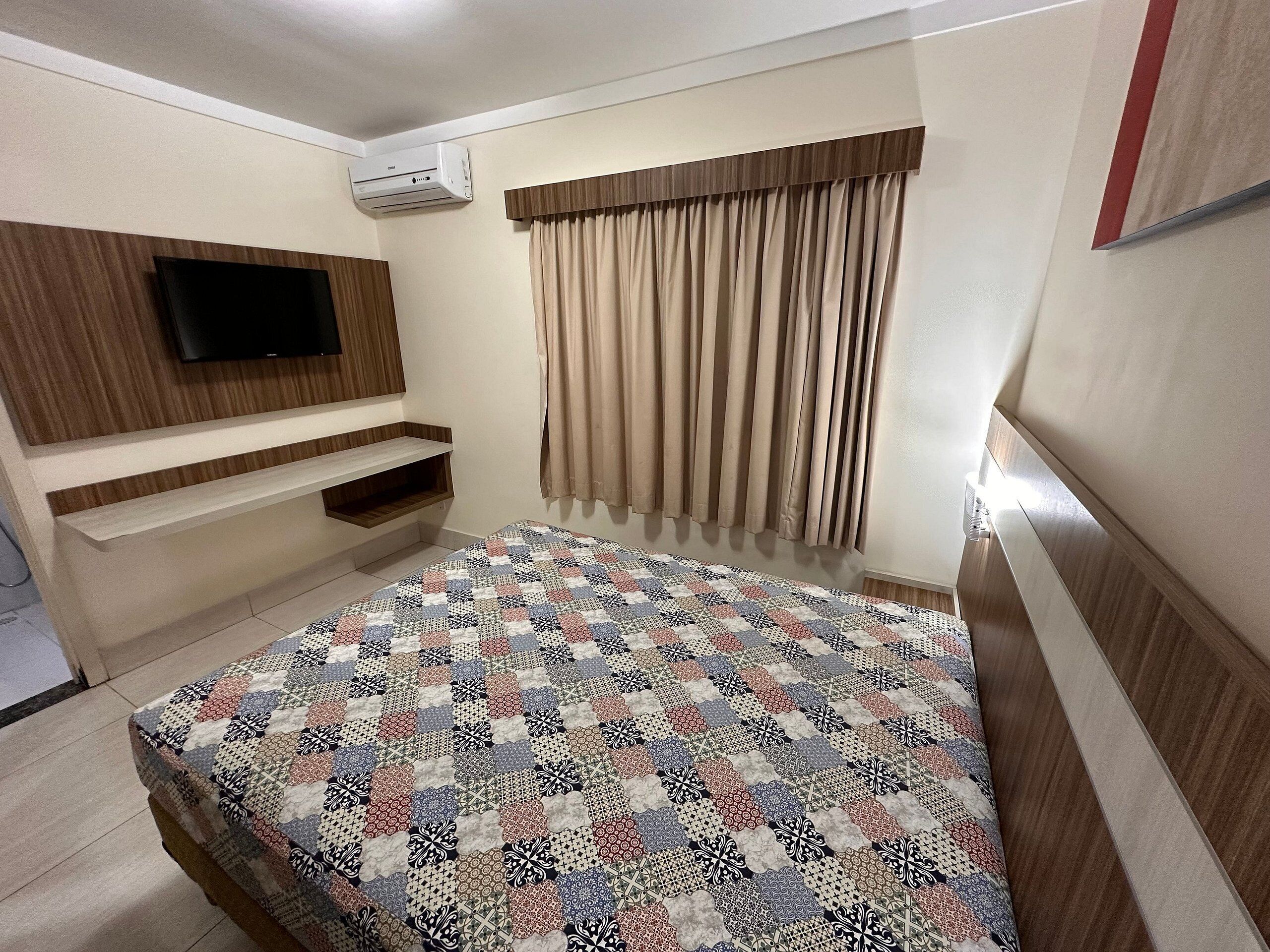 A hotel room with a bed, a TV mounted on the wall, a window with curtains, and a built-in shelf.
