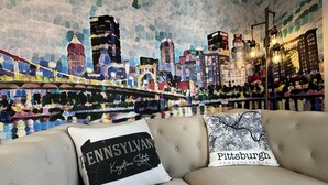 A room with a cityscape mural on the wall, a beige sofa, and two decorative pillows with text. - Hidden Pearl - City Skyline View - Free Hot Spa (Pittsburgh)