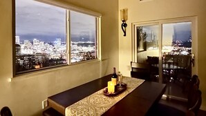 A dining area with a table set for two, a candle, and a bottle of wine. There are multiple windows offering a city view. - Hidden Pearl - City Skyline View - Free Hot Spa (Pittsburgh)