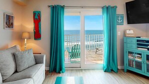 Une pièce comprenant un canapé, une petite table avec une lampe, un meuble turquoise et une vue sur la plage, accessible par une porte coulissante.