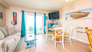 Un salon comprenant un canapé, une table basse et un coin repas avec des chaises et une table. Un téléviseur est fixé au mur et un tableau représente un bateau.