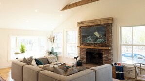 A living room with a brick fireplace, a large window, and a sofa with cushions. - Katama Retreat W Pickle ball Court (Edgartown)