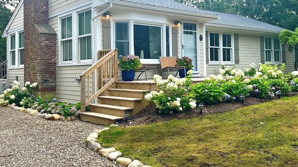 A two-story house with a covered porch, a brick chimney, and a garden with white flowers. - Katama Retreat W Pickle ball Court (Edgartown)