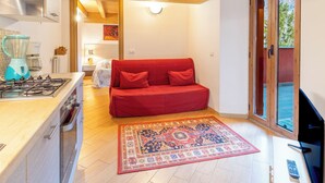 Une cuisine compacte équipée d’une cuisinière à gaz, d’un mixeur et d’une machine à café. Le salon comprend un canapé rouge avec des coussins et un tapis à motifs sur le sol.