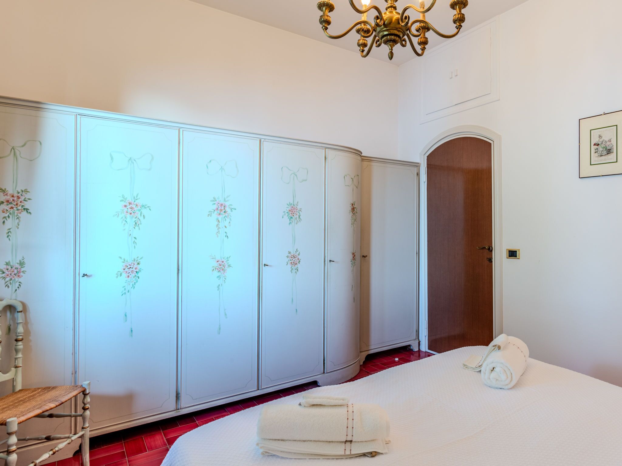A bedroom with a wardrobe, a bed, a chair, and a chandelier.
