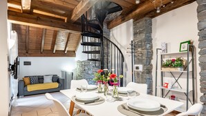 Ein Essbereich mit einem weißen Tisch, gedeckt für vier Personen, umgeben von weißen Stühlen. Im Hintergrund ist eine schwarze Wendeltreppe zu einer oberen Ebene hin sichtbar.