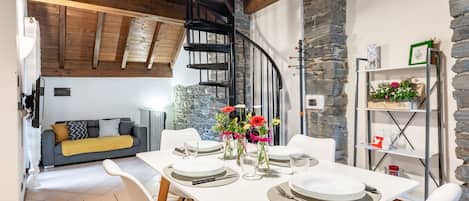 A dining area with a white table set for four, surrounded by white chairs. A black spiral staircase is visible in the background, leading to an upper level.