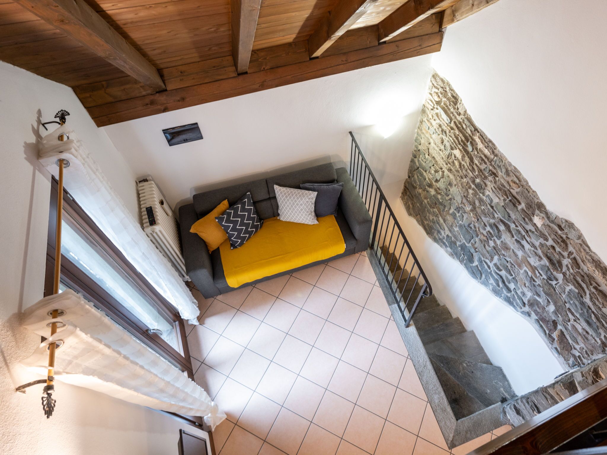 Habitación con techos altos, un sofá gris, manta amarilla y almohadones estampados. Se ve una pared de piedra y una escalera.