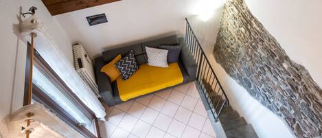 Habitación con techos altos, un sofá gris, manta amarilla y almohadones estampados. Se ve una pared de piedra y una escalera.
