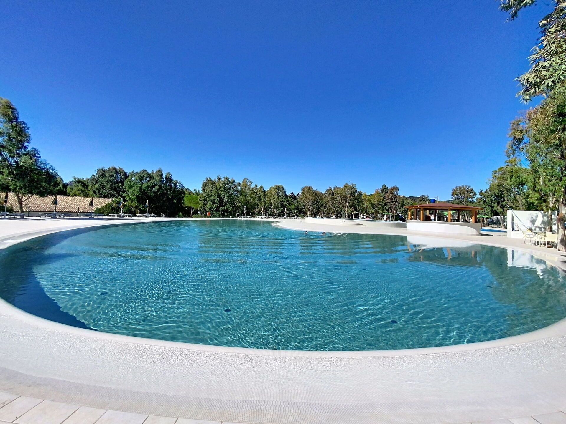 A large, clear swimming pool with a sandy beach area, surrounded by trees and a pavilion.
