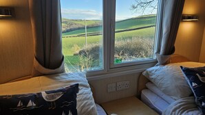 A bedroom with a large window offering a view of rolling hills. There are two beds with pillows and a bedside table. - Penmor -  a holiday home that sleeps 6 guests  in 3 bedrooms (Bude)