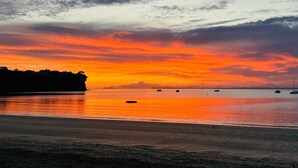 A beach at sunset with boats on the water and a silhouette of a cliff. - Stay and Relax at Beachfront Beauty (Whangaparāoa)