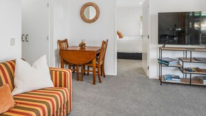 A modern, well-lit room with a dining table, chairs, a striped sofa, a TV, and a bookshelf. - Stay and Relax at Beachfront Beauty (Whangaparāoa)