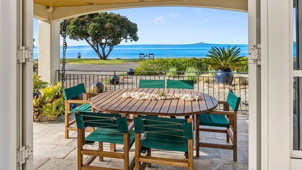 An outdoor patio with a wooden table and chairs, a view of the ocean, and a large tree. - Stay and Relax at Beachfront Beauty (Whangaparāoa)